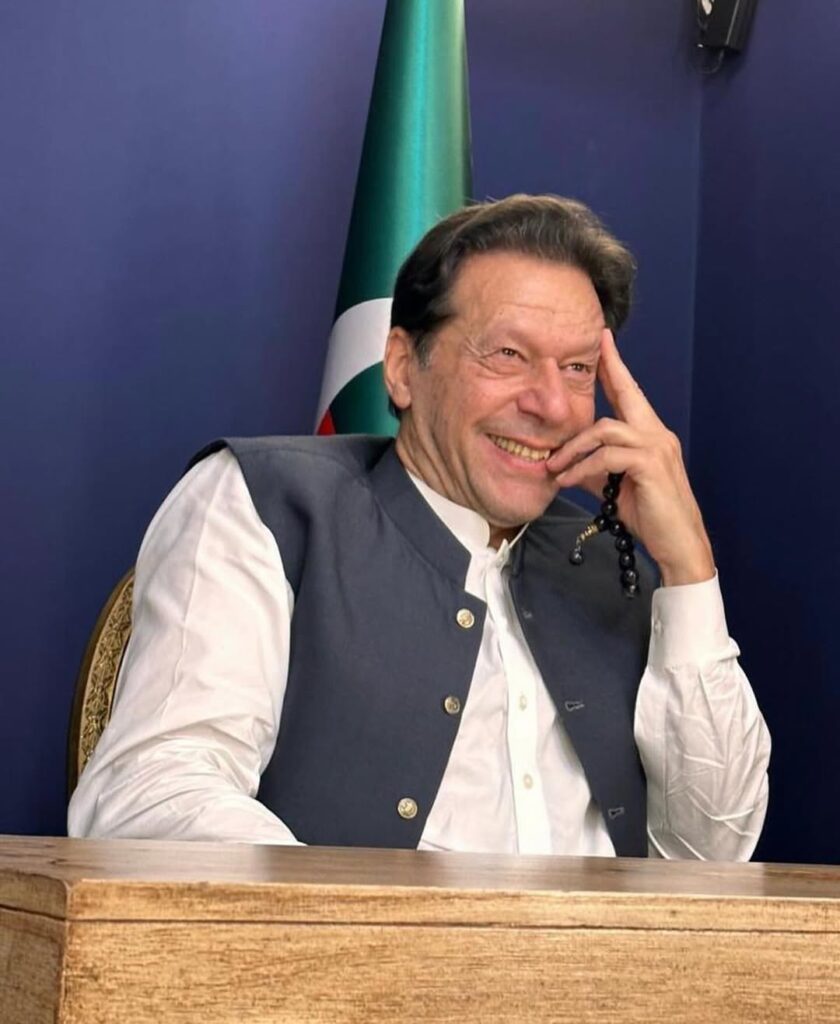 Smiling man in a white shirt and gray vest sits at a wooden desk, holding prayer beads. A green flag is visible in the background. Warm and relaxed atmosphere. Imran khan